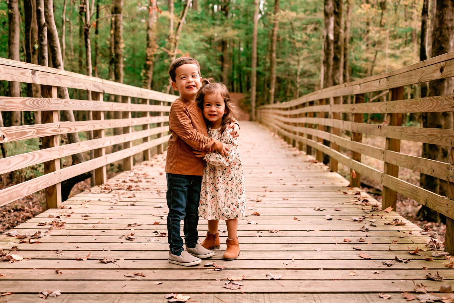 A young boy and girl share a sweet hug while standing on a wooden bridge surrounded by fall foliage.