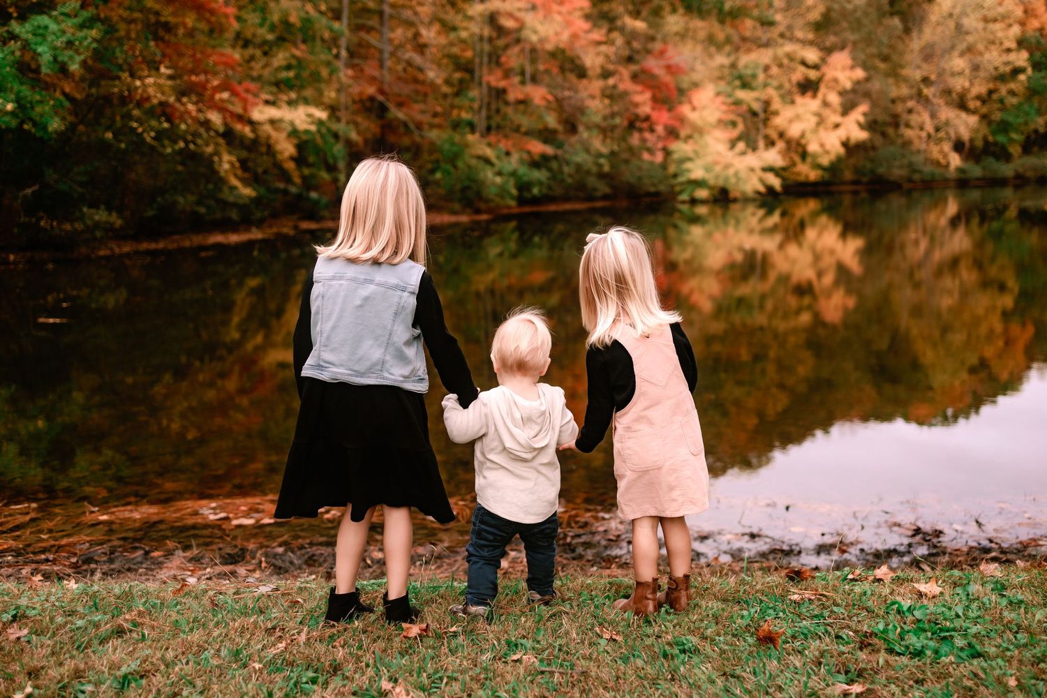 Three young siblings hold hands while gazing at a serene autumn pond surrounded by colorful fall trees.