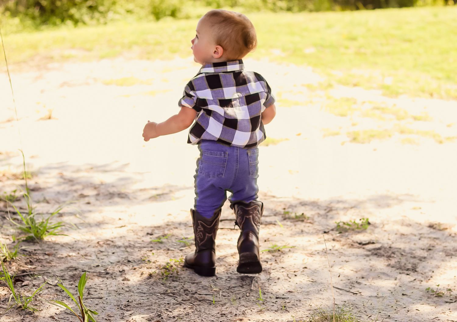 Toddler boy in a plaid shirt and boots walking on sandy ground outdoors.