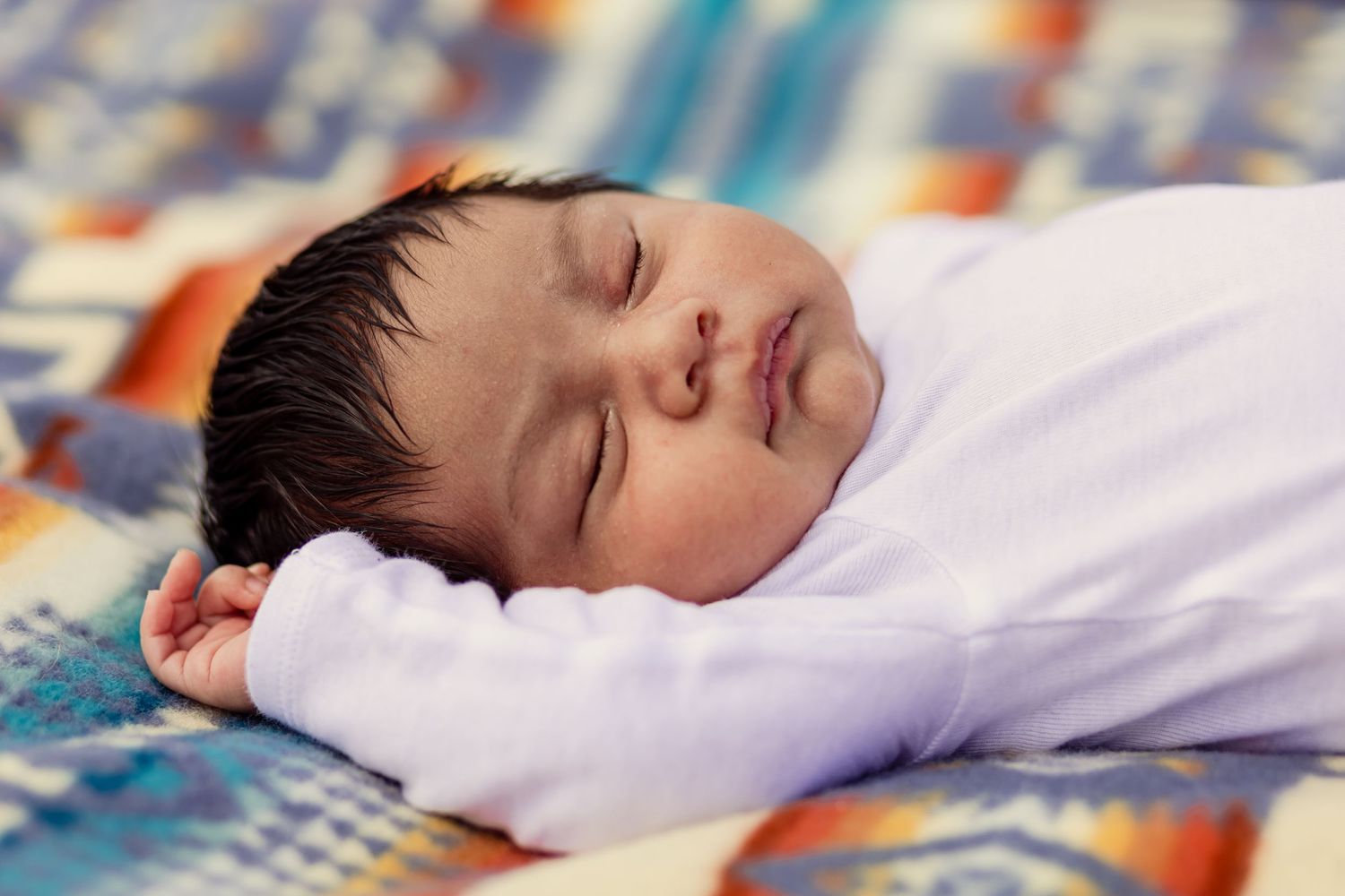 Peaceful sleeping newborn baby with dark hair resting on colorful patterned blanket in white onesie.