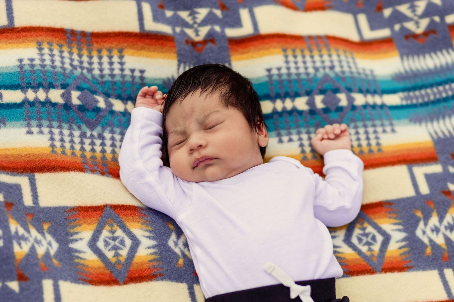 Sleeping newborn baby in white onesie with arms raised lying on colorful Pendleton patterned blanket.