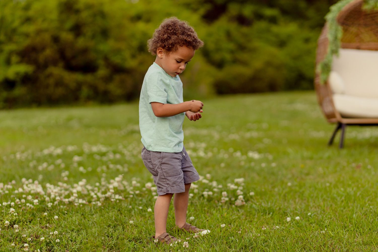 Toddler boy in mint shirt and gray shorts stands barefoot on green grass examining small object in hands.