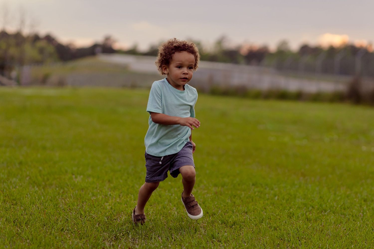 Young toddler boy in mint shirt and gray shorts runs across green grass field at dusk outdoors.
