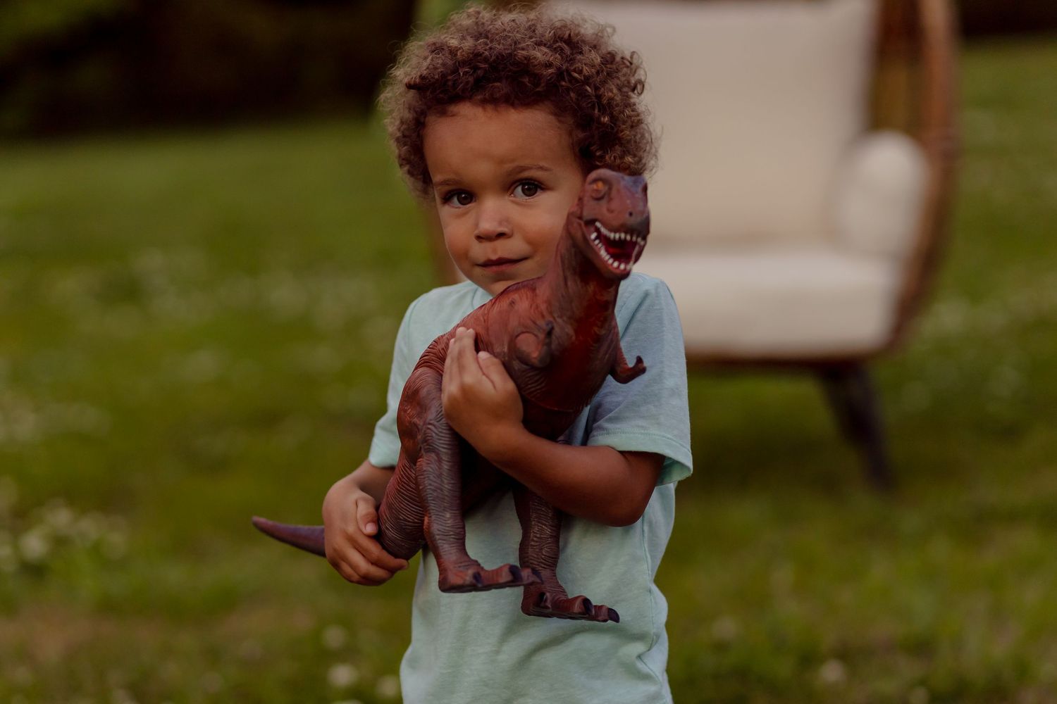 Young boy with curly hair hugs large brown T-Rex dinosaur toy while standing on green grass outdoors.