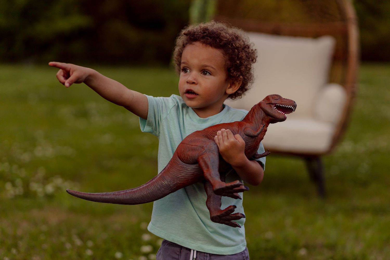 Curly-haired toddler boy in mint shirt holds brown dinosaur toy while pointing in lush green backyard.