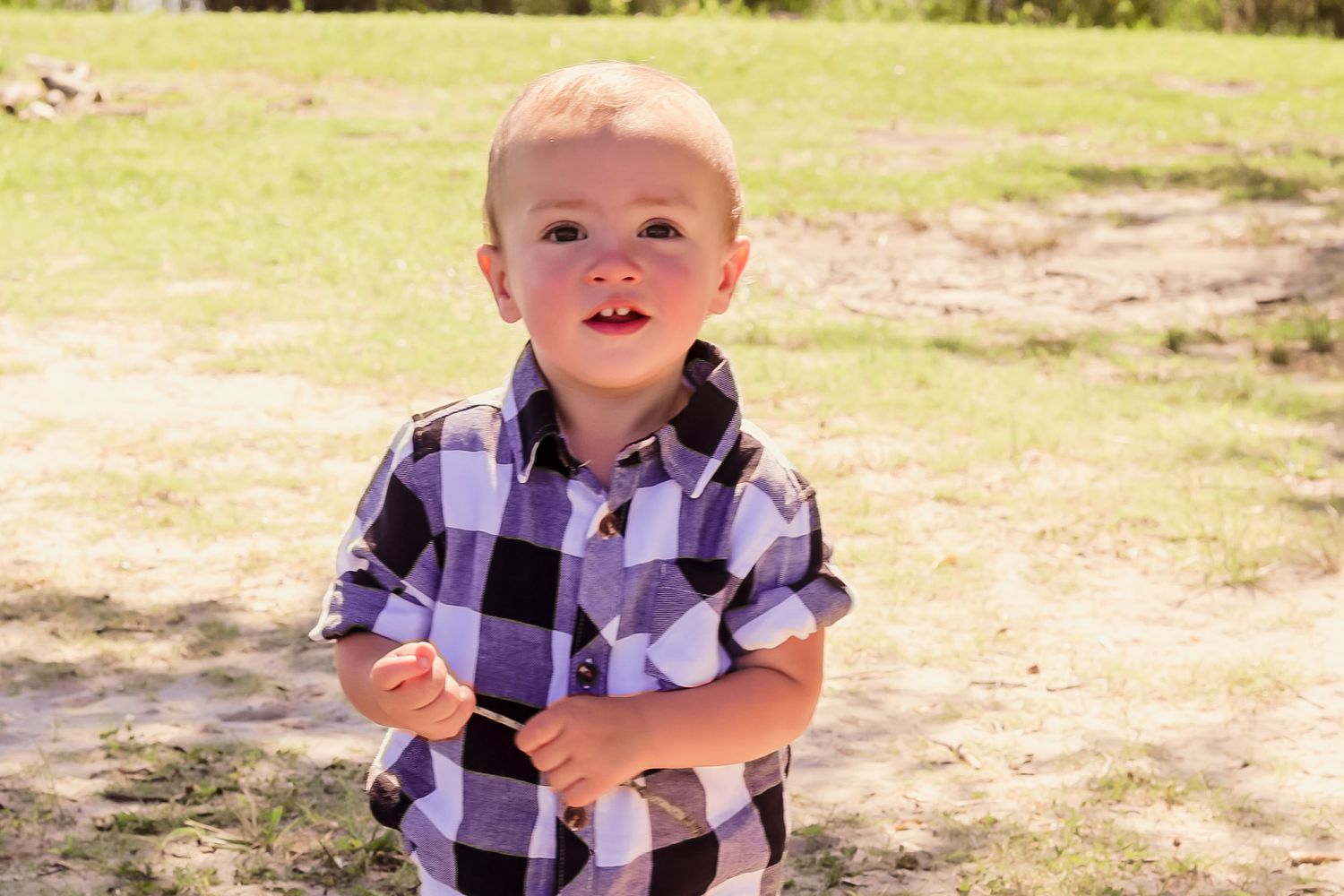 Blonde toddler boy in plaid shirt standing outdoors in a sunny park.