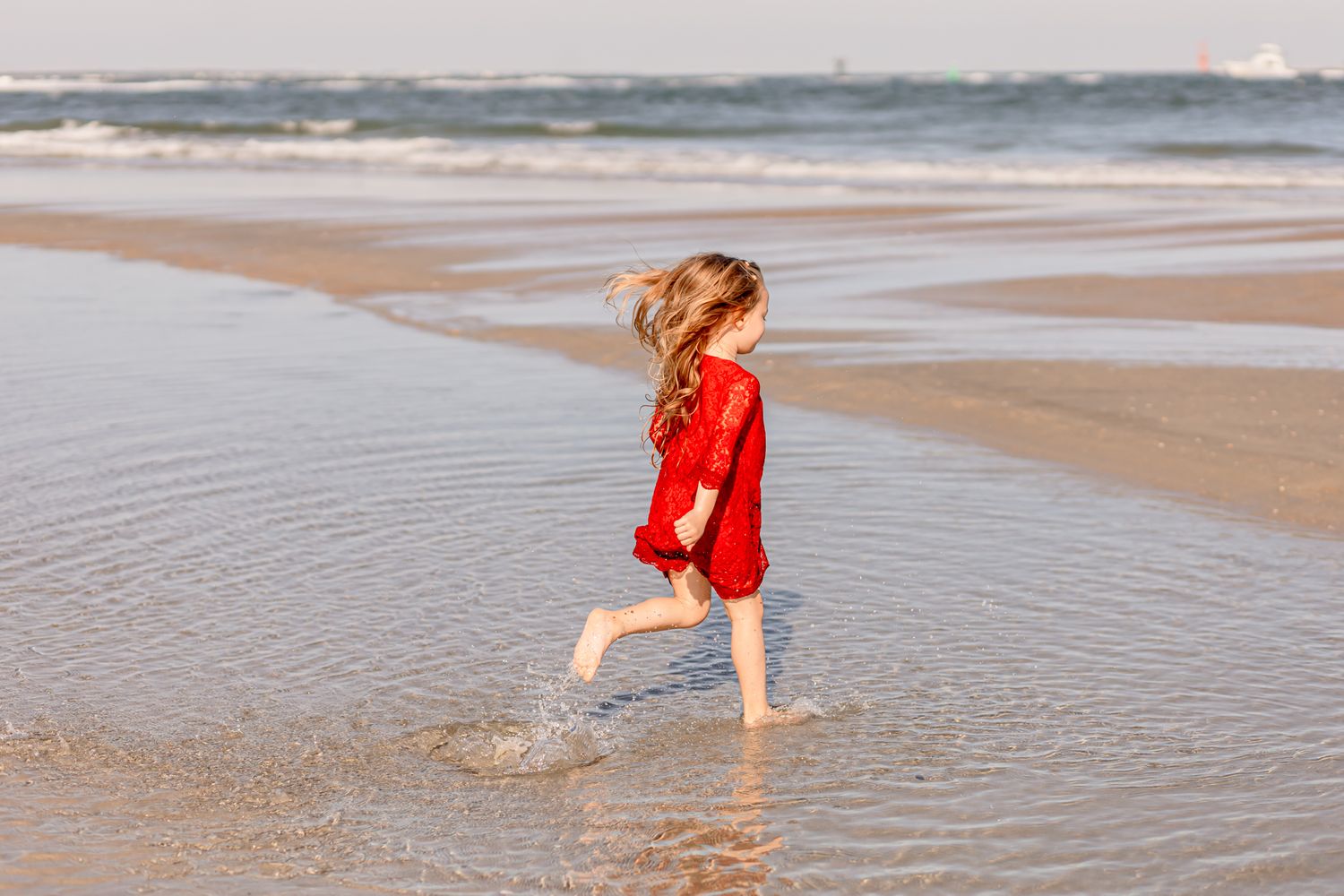 Young girl in red dress running barefoot along the shoreline on a sunny beach.