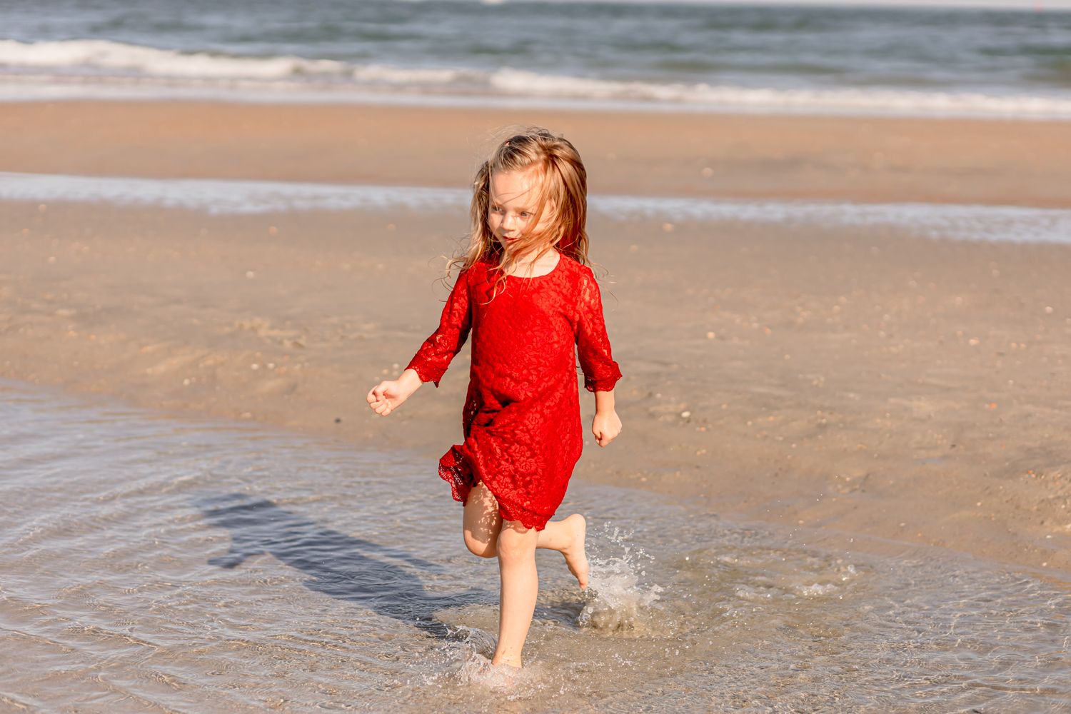 Little girl in red lace dress splashing through shallow ocean waves on a sandy beach.