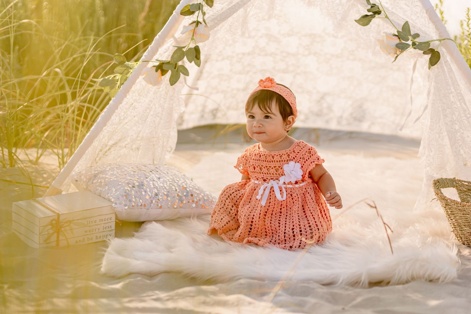 Adorable baby girl in peach crochet dress sitting near a white lace teepee on a fur rug.