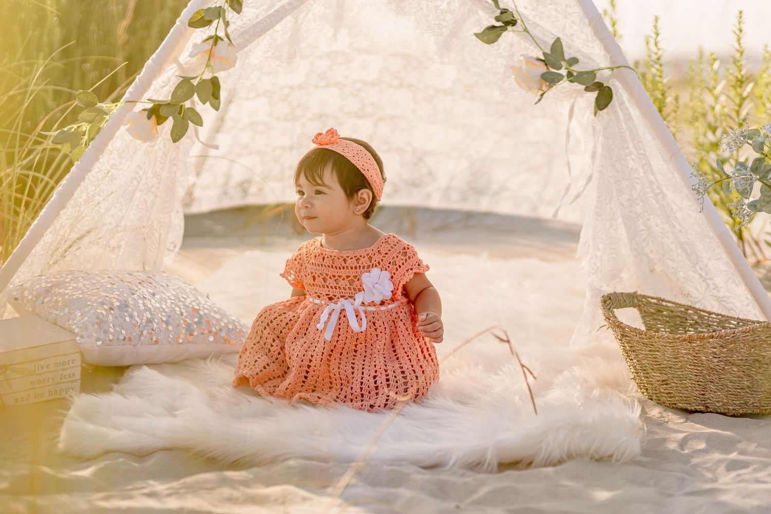 Sweet baby girl in orange knit dress sitting peacefully inside a white lace teepee outdoors.