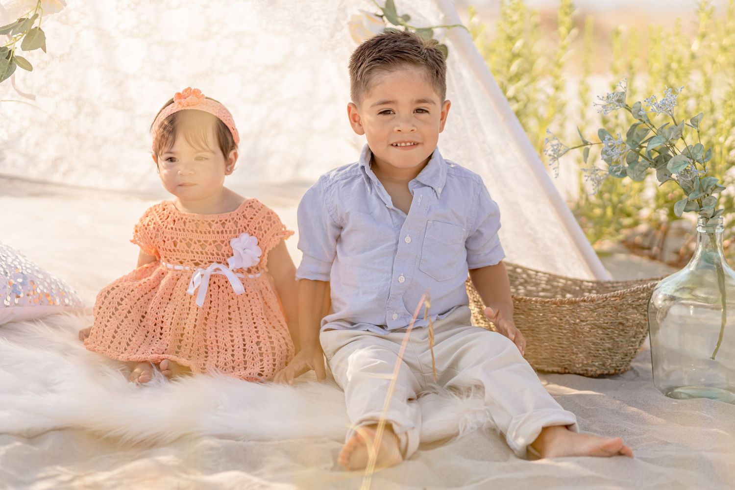 Cute baby girl in peach crochet dress and young boy in blue shirt posing by a lace teepee.