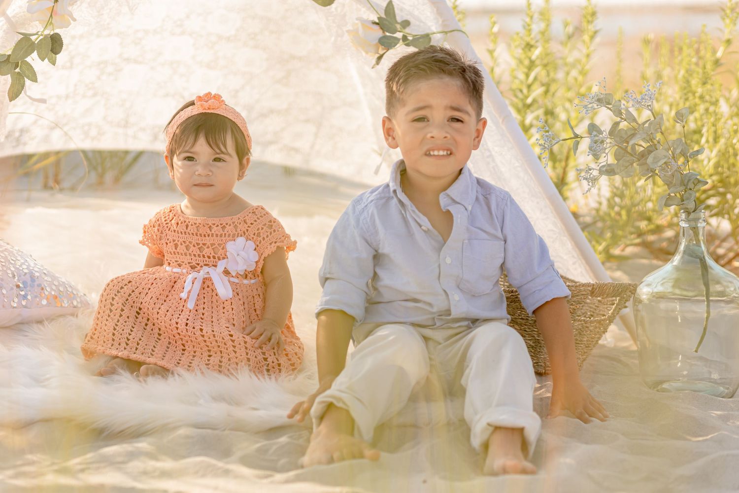 Two young siblings smiling together in coordinating outfits beside a white lace teepee outdoors.