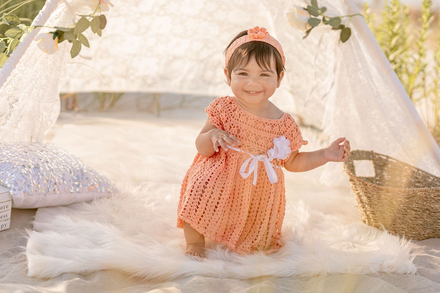 Happy baby girl in peach crochet dress smiling and standing near a wicker basket by a teepee.