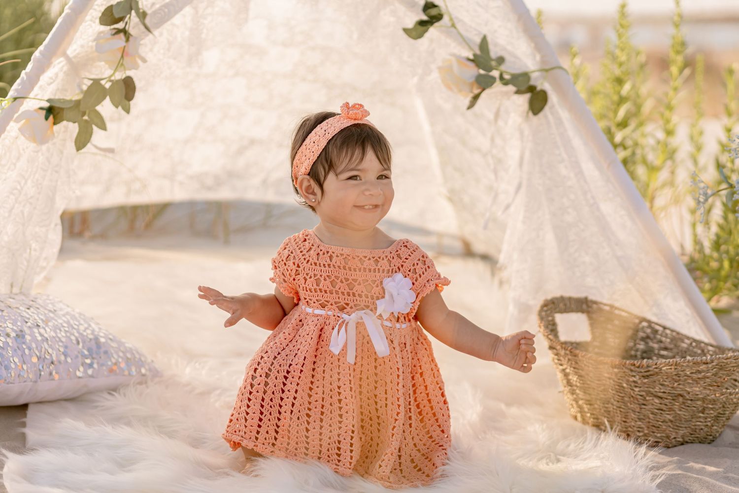 Joyful baby girl in orange knit dress with headband laughing beside a white lace teepee.