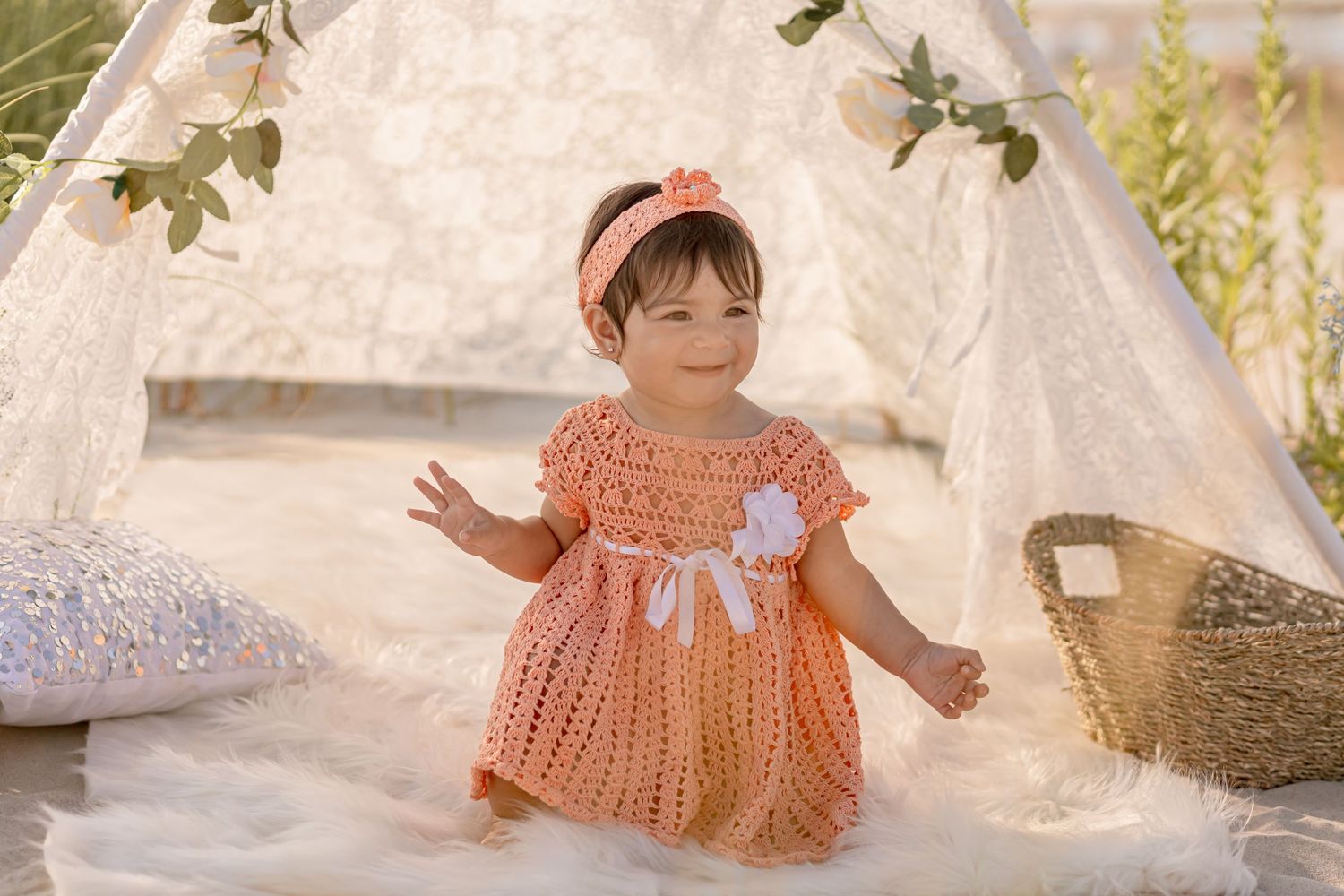 Smiling baby girl in peach knit dress waving beside a white lace teepee on a fluffy rug.