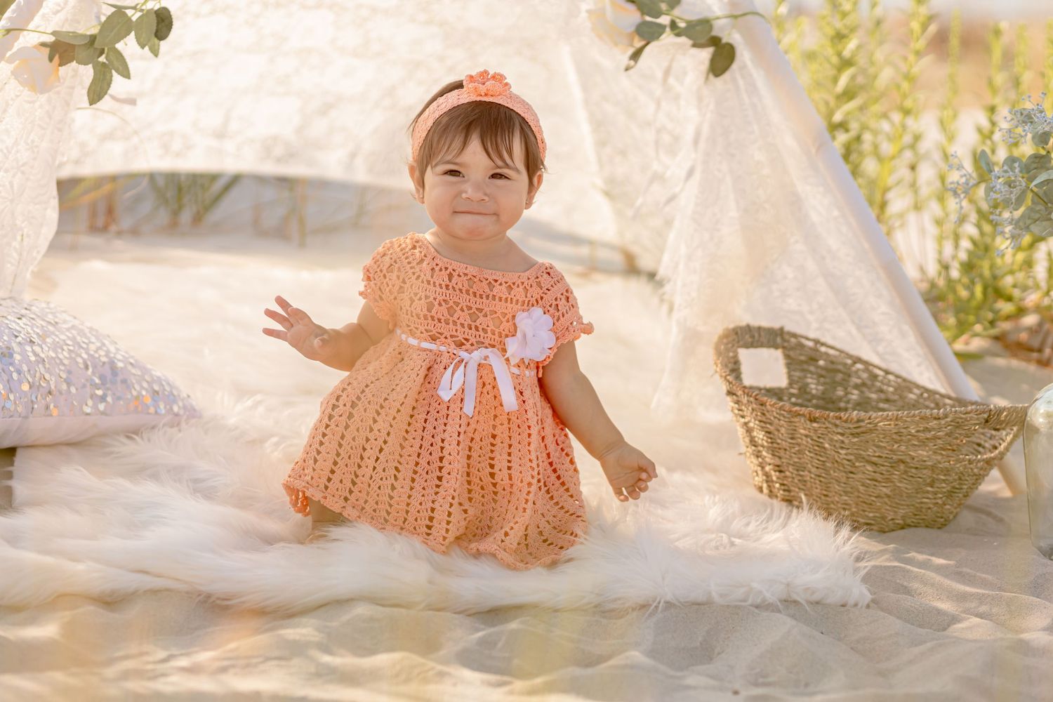 Cheerful baby girl in peach crochet dress sitting on fur rug near a lace teepee and basket.