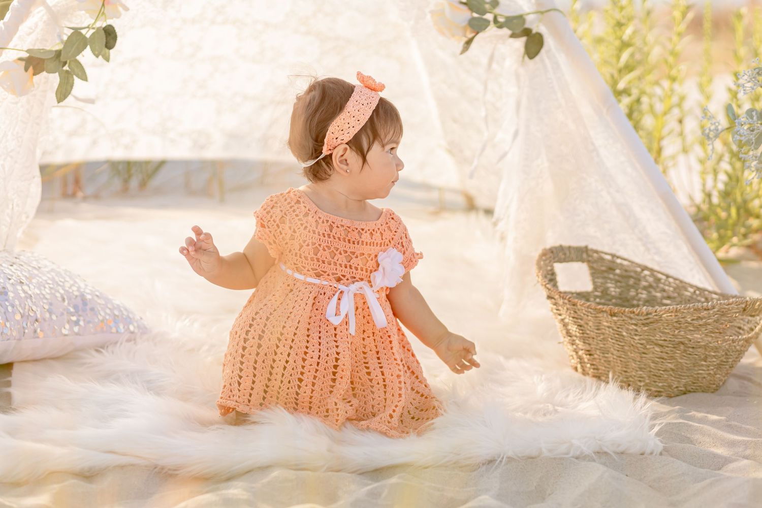Baby girl in peach crochet dress and headband sitting on white fur rug near decorative teepee.