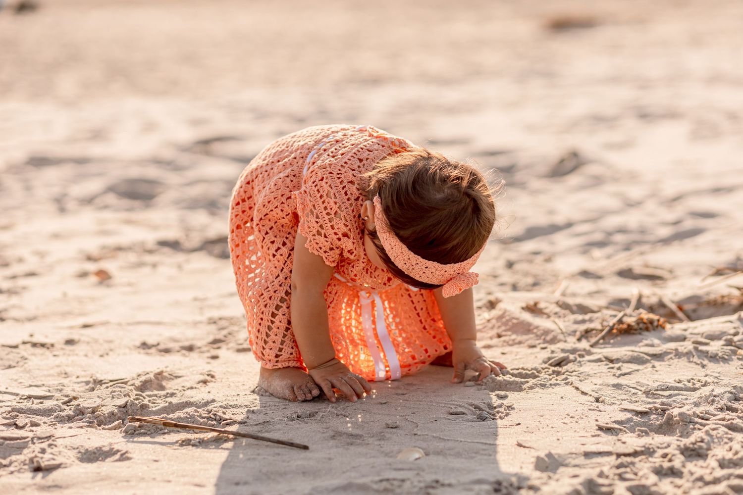 Toddler girl in orange crochet dress bending down to explore sandy beach.