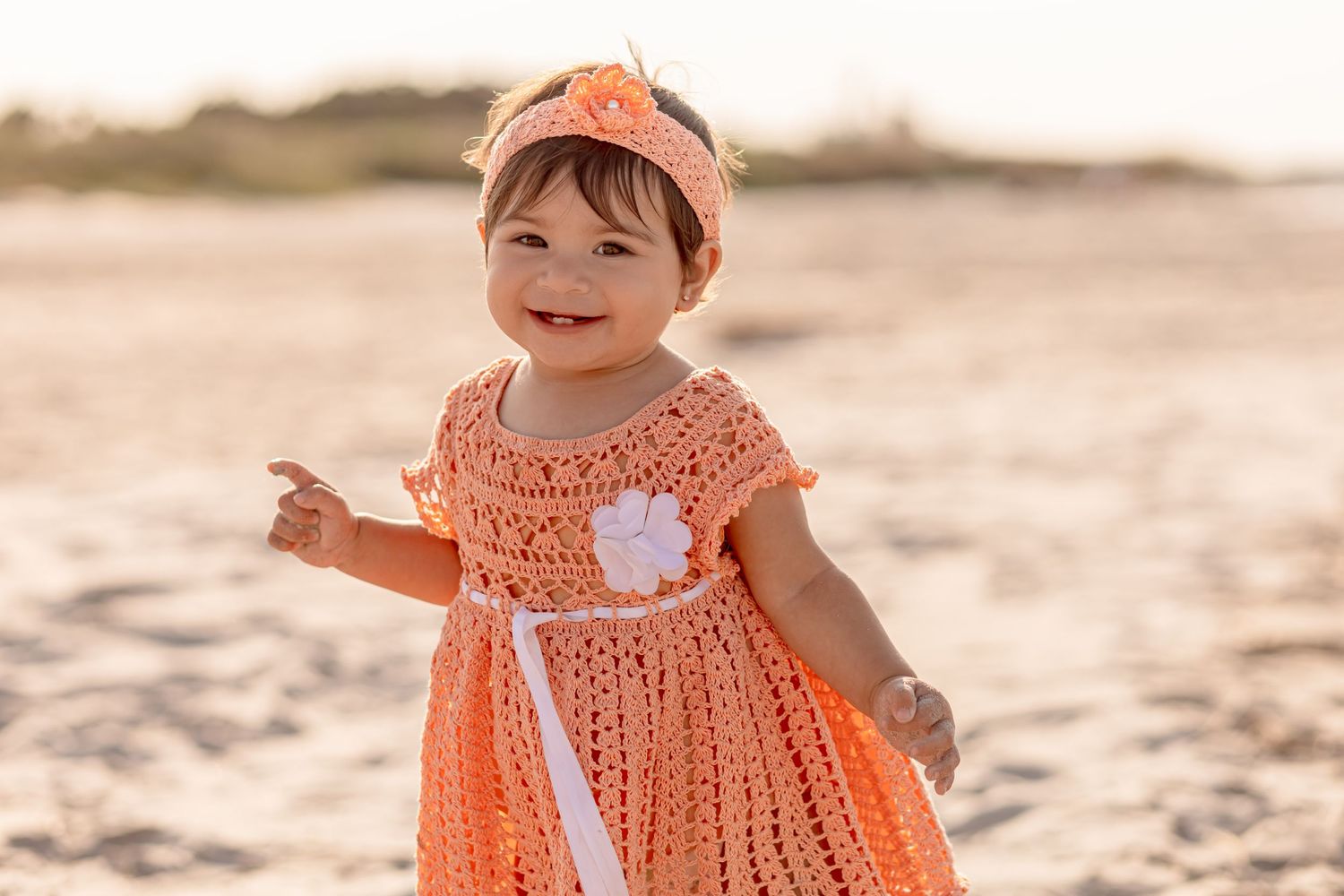 Happy toddler girl in orange crochet dress with flower detail smiling on sunny beach.