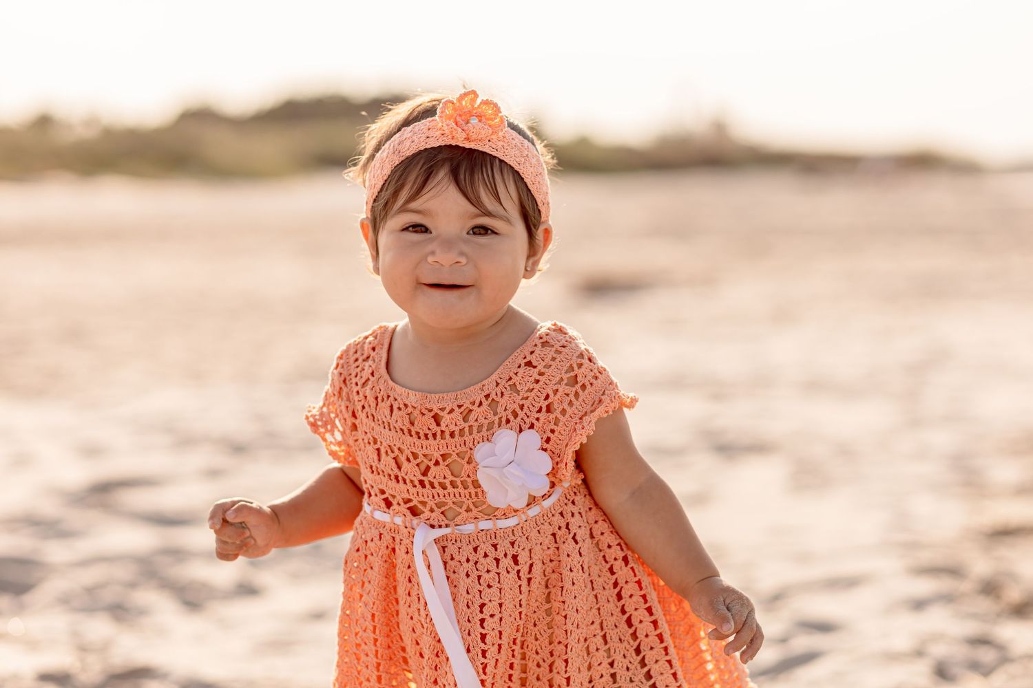 Smiling baby girl in peach crochet dress and floral headband standing on sandy beach.