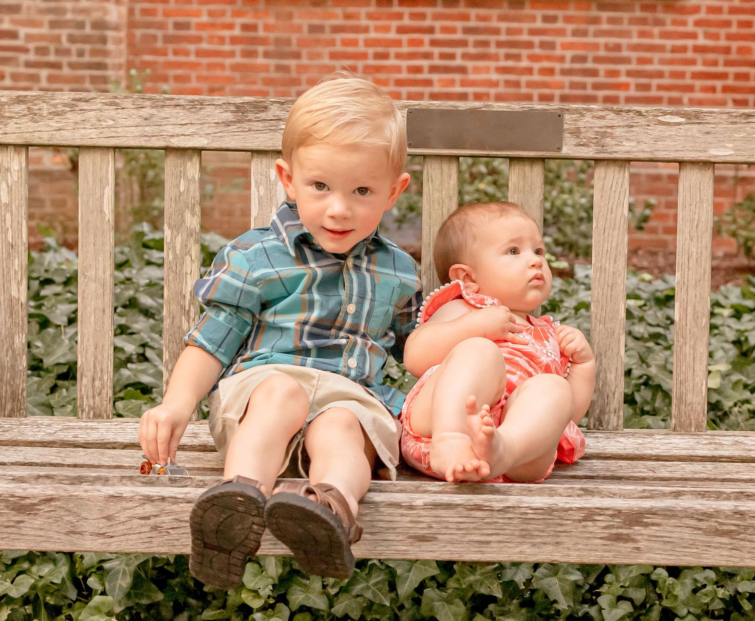 A young blonde boy in a plaid shirt sits on a wooden bench beside an infant in a coral outfit.