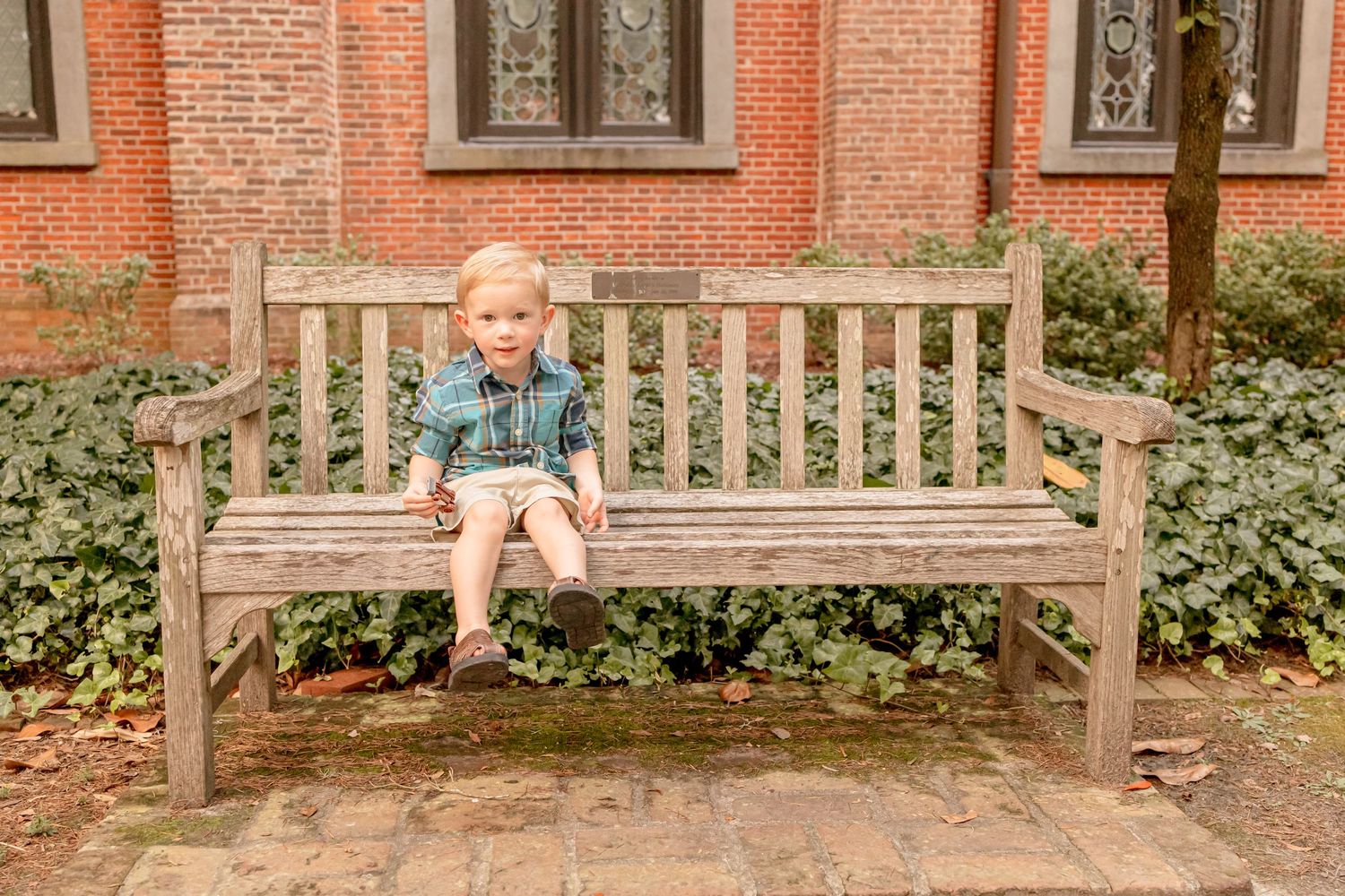 A blonde toddler boy in a denim shirt sits alone on a large wooden bench near a brick building.