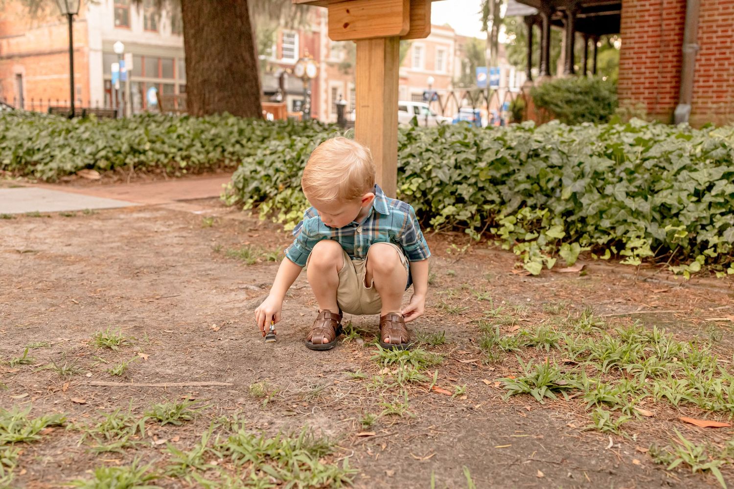 A young blonde boy in a plaid shirt crouches down examining something on the ground outdoors.