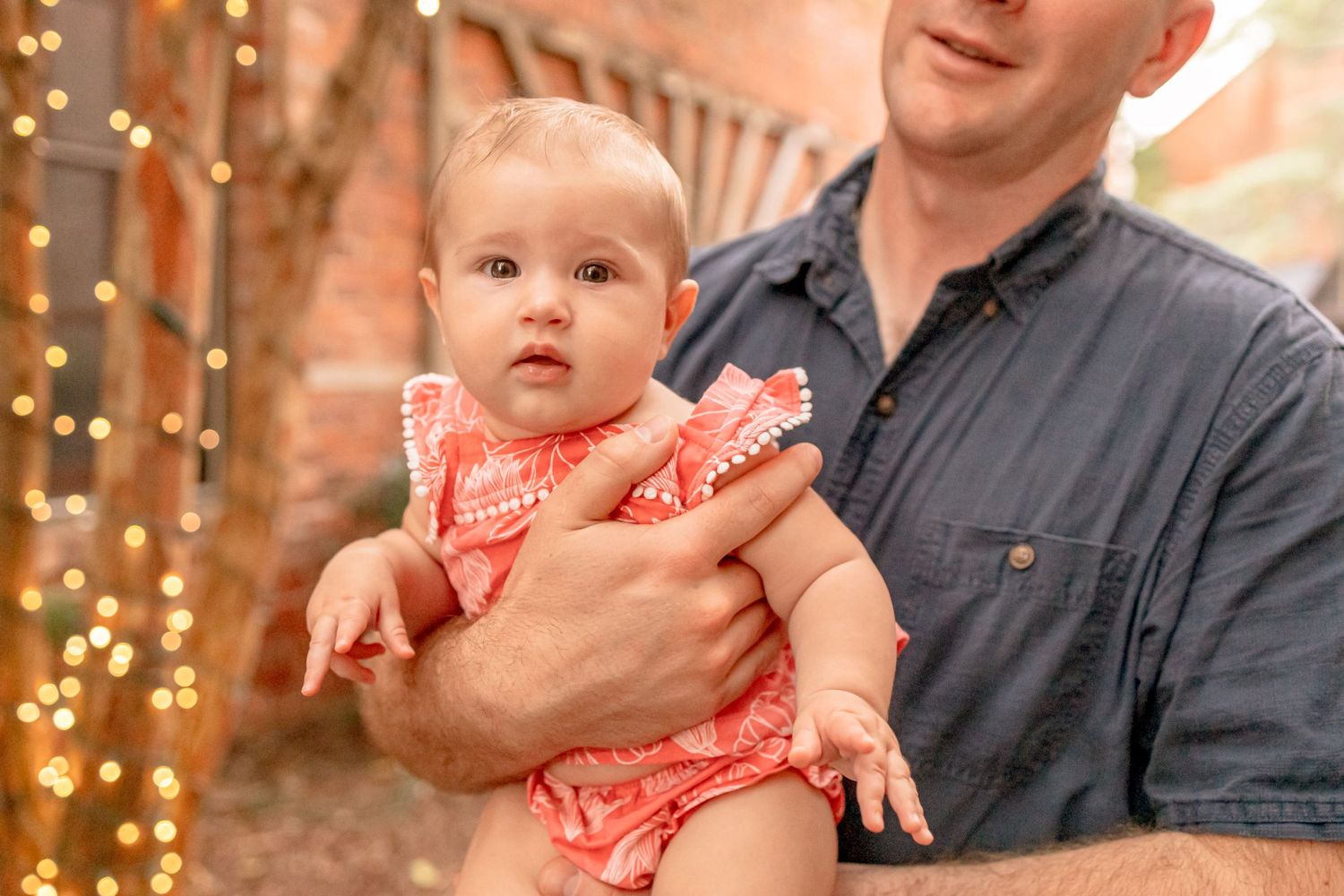 A man in a denim shirt holds a baby girl in a coral romper near twinkling string lights outdoors.