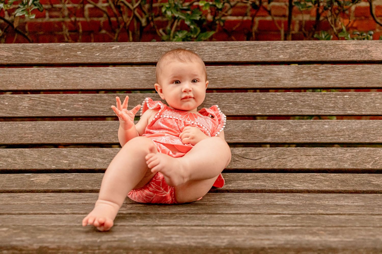 A happy baby girl in a coral romper sits on a wooden bench waving her hand at the camera.