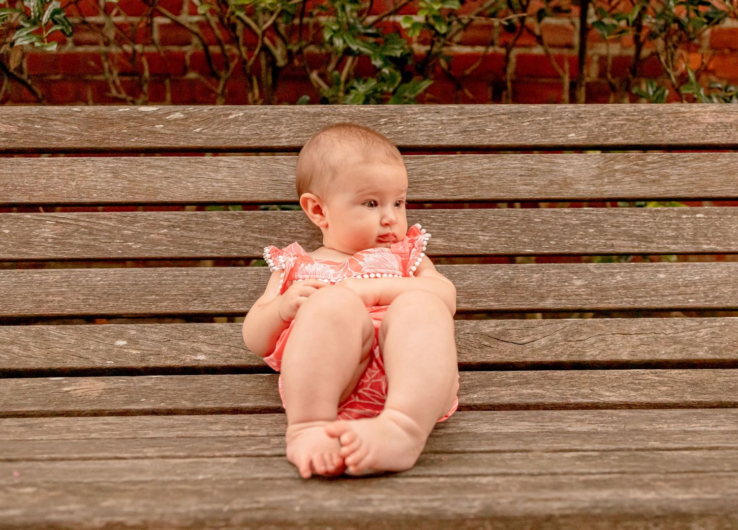 A baby girl in a coral romper sits propped against a wooden bench slat outdoors in a garden.
