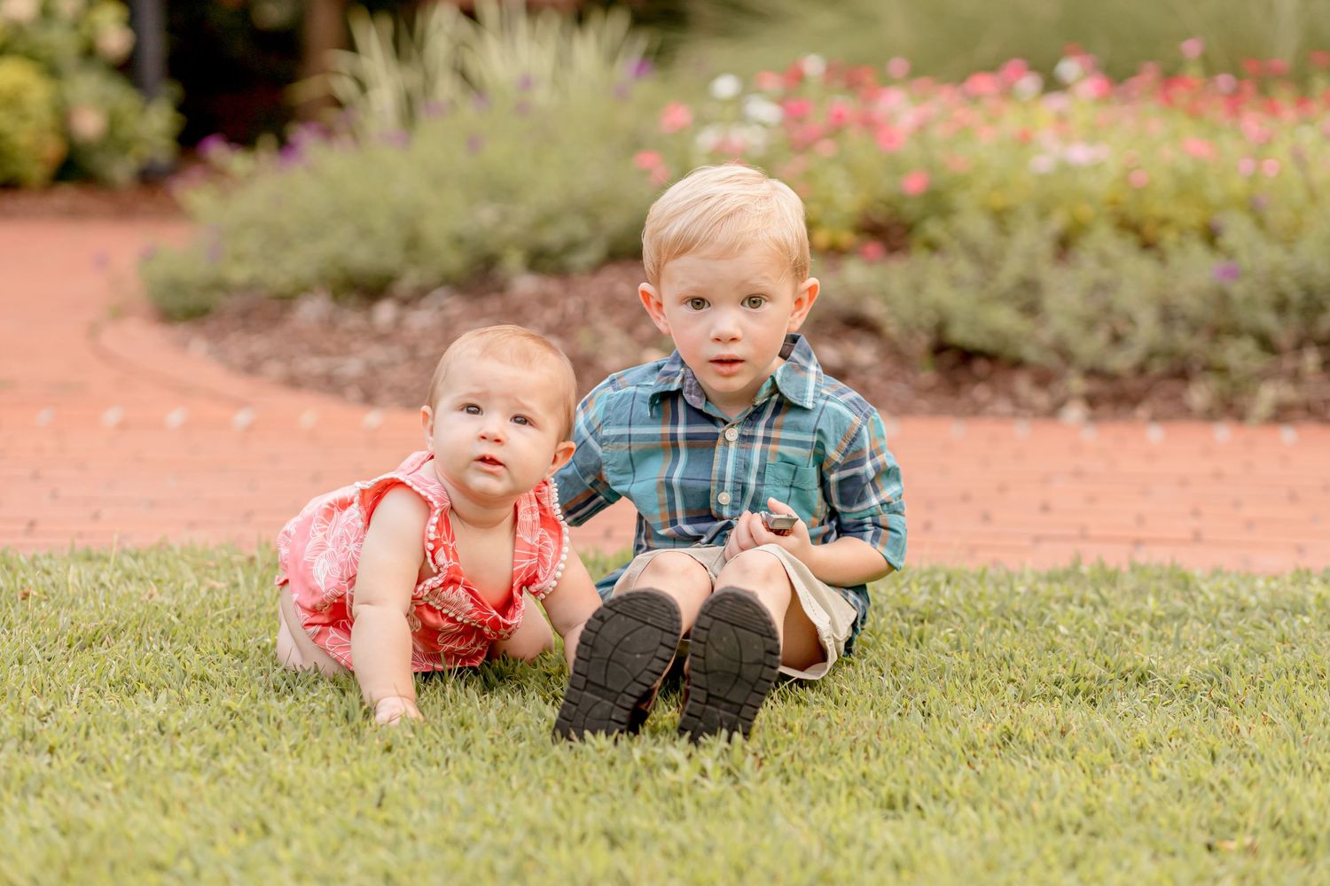 Two young siblings, a baby girl and toddler boy, sitting together on green grass outdoors.