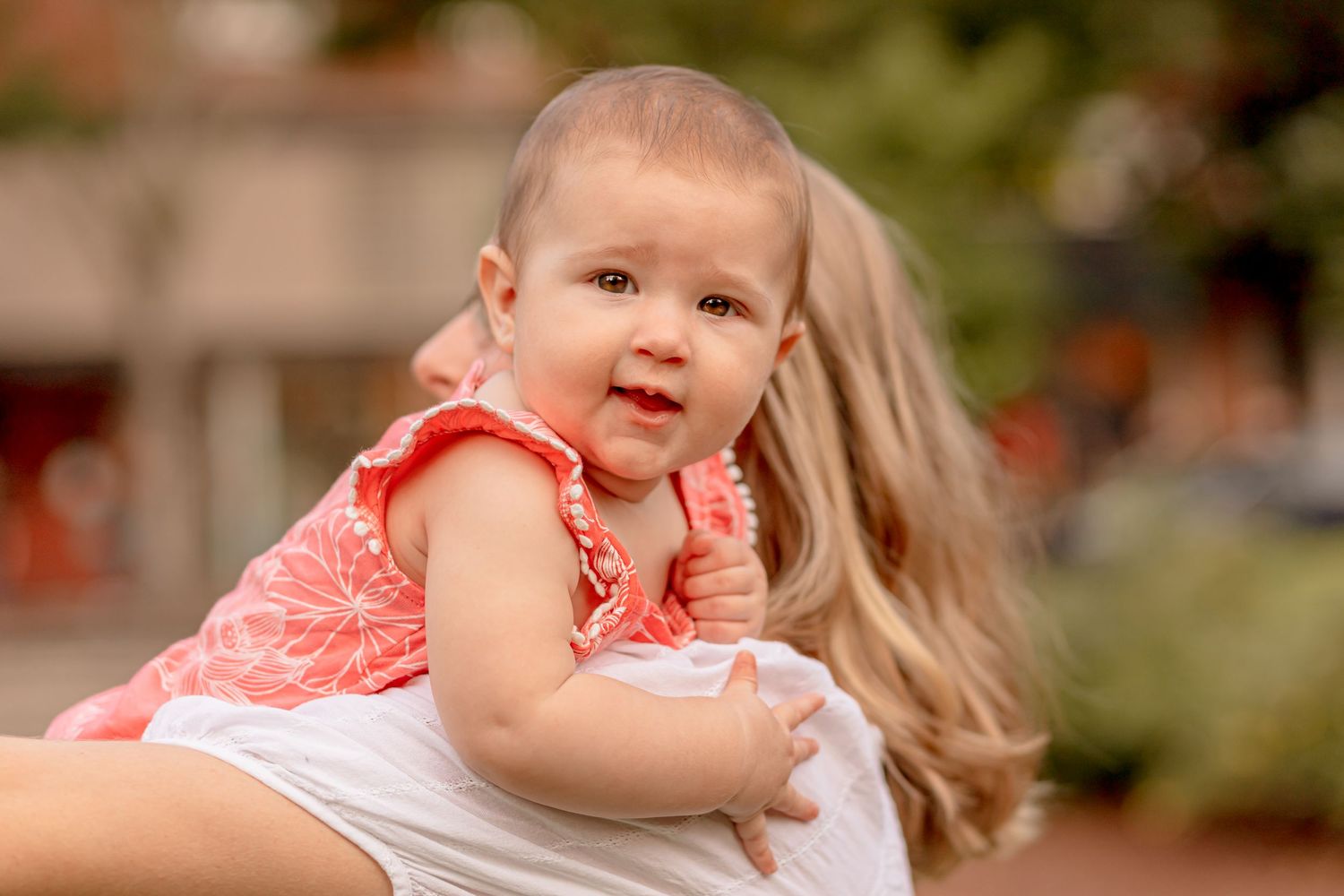 Happy baby girl in pink floral dress being held by a blonde woman outdoors.