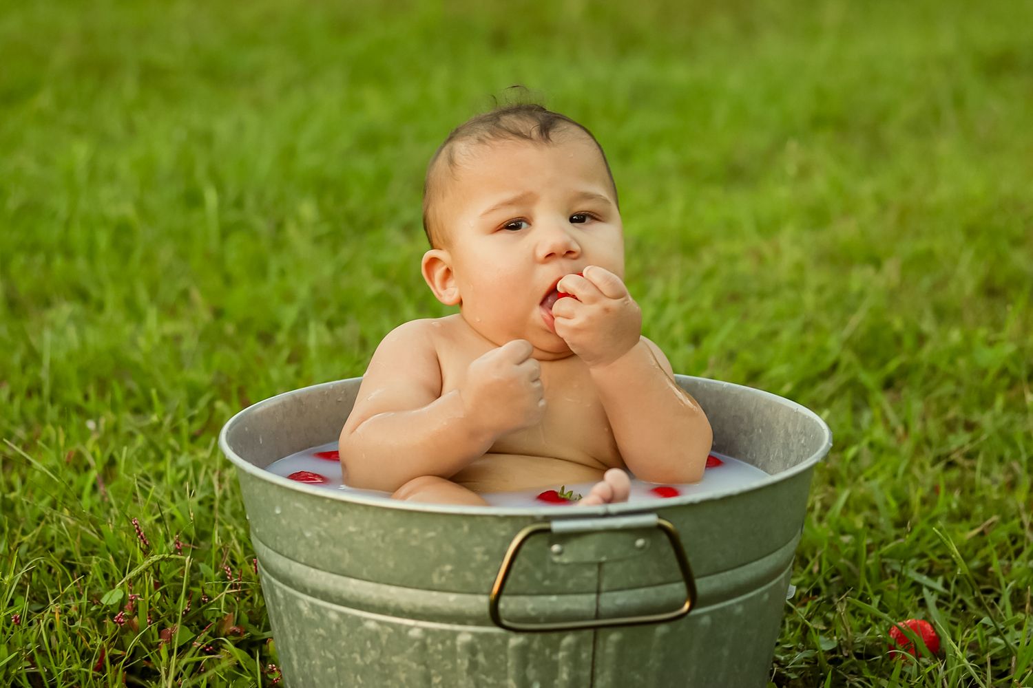 Baby sitting in a galvanized tub outdoors on green grass eating strawberries.