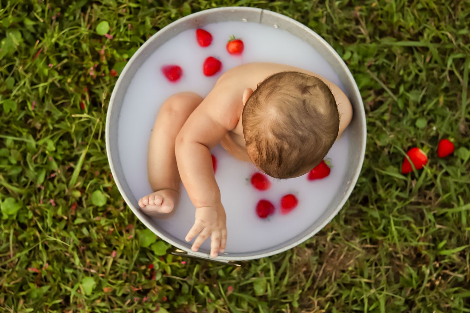 Baby relaxing in a round tub of milk with red strawberries on green grass.
