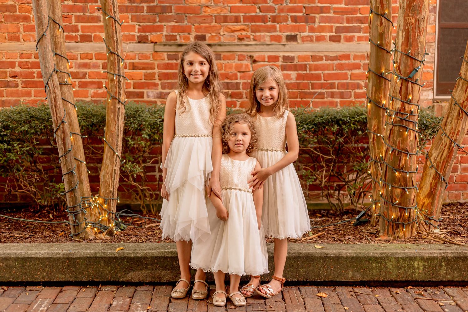 Three sisters in white dresses posing together near brick wall with string lights.
