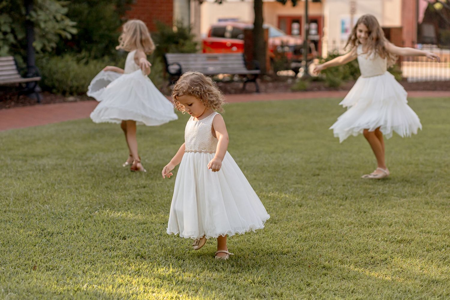Three girls in white dresses twirling and spinning on green grass in outdoor setting.