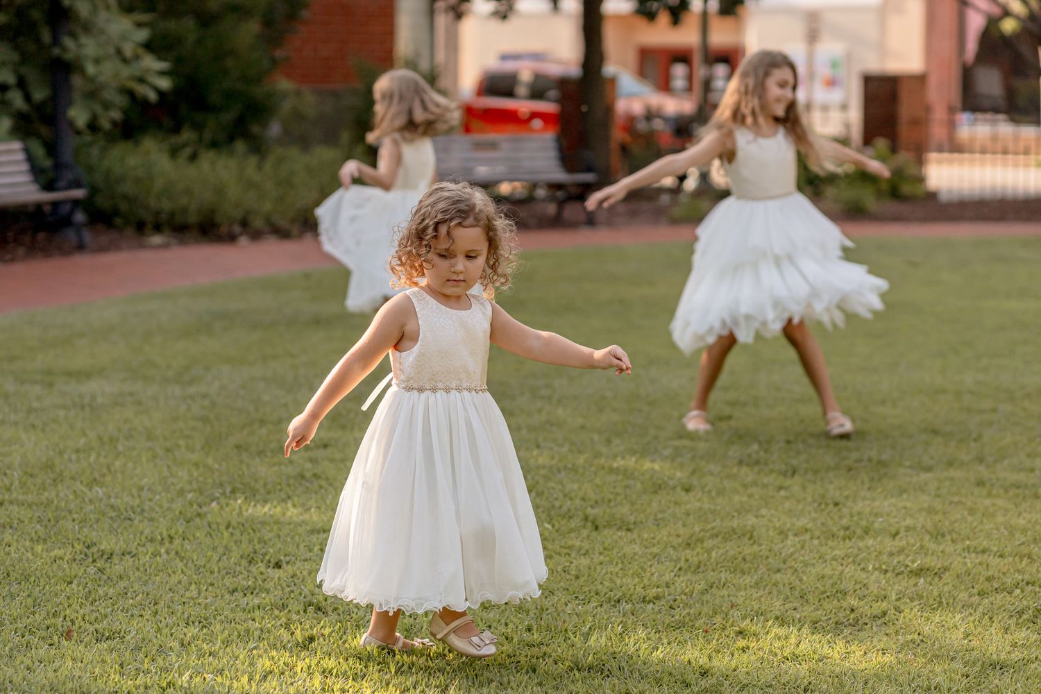 Three girls in white dresses dancing and spinning on green lawn at golden hour.