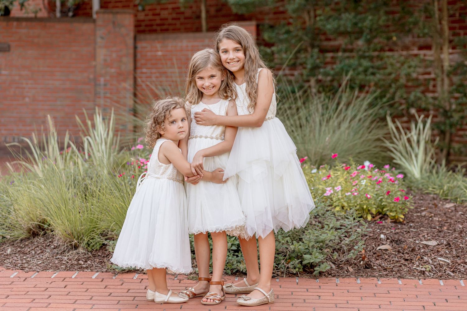 Three young girls in white dresses hugging each other on brick pathway in garden.
