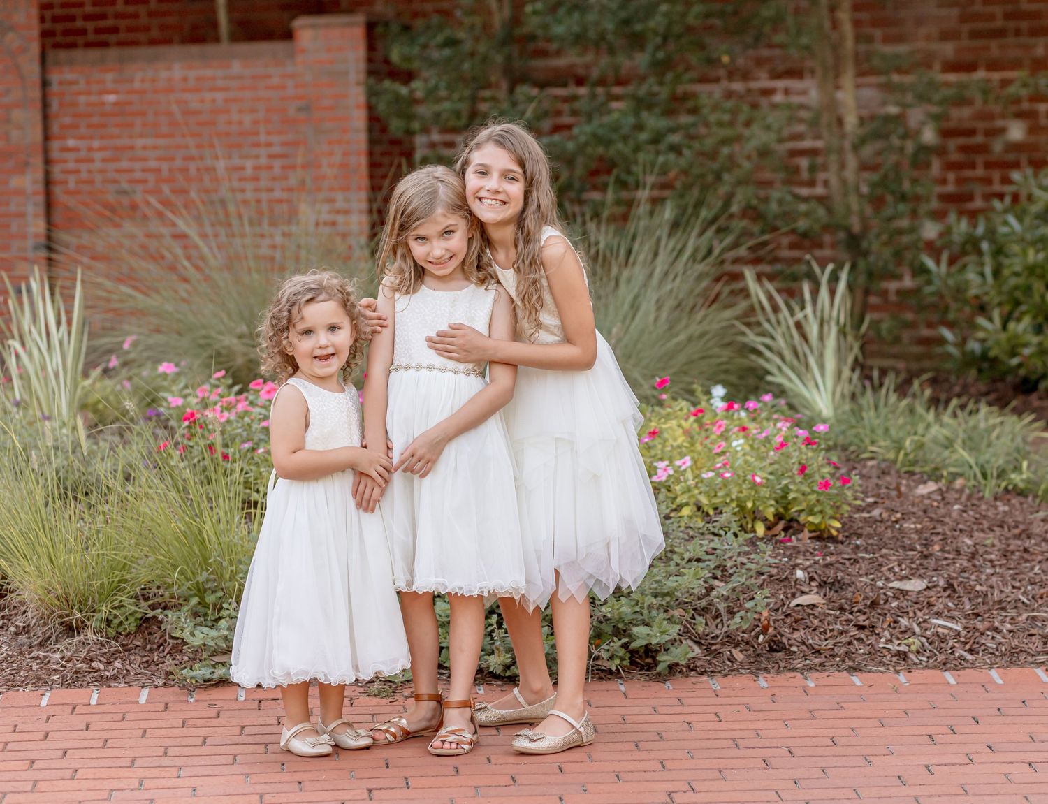 Three smiling sisters in white dresses posing together on brick path with garden backdrop.