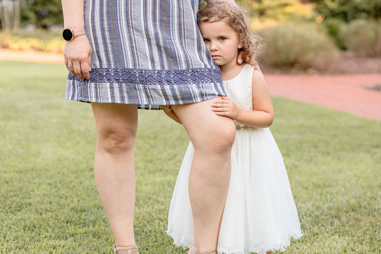 A toddler girl in a white dress clings shyly to her mothers leg outdoors on a green lawn.