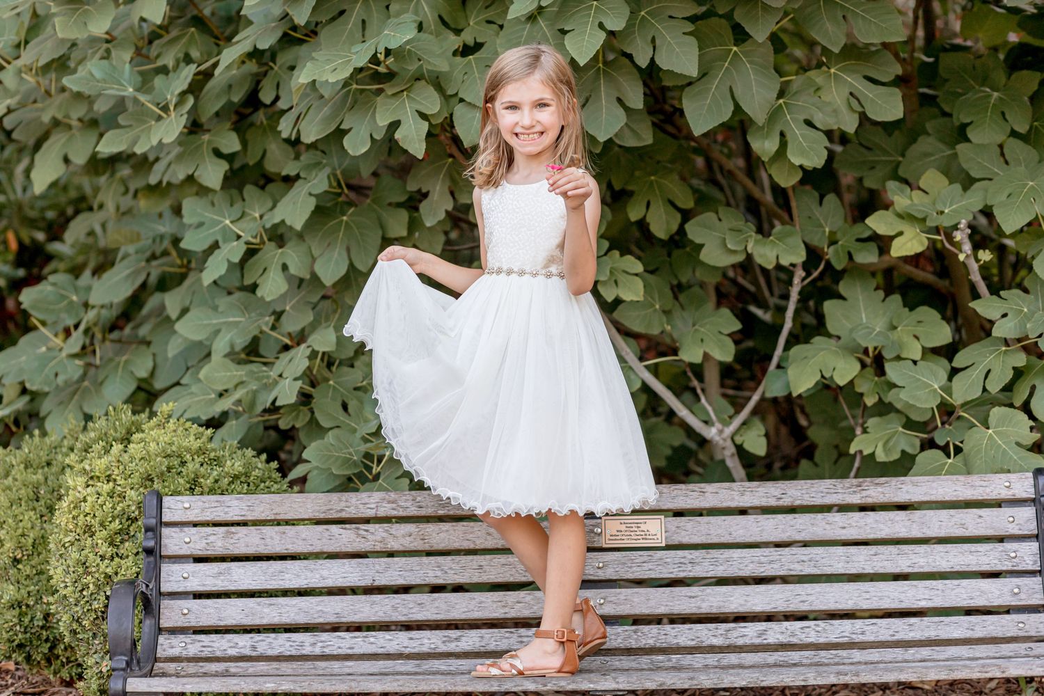 A young girl in a white flower girl dress twirls on a wooden bench surrounded by lush greenery.