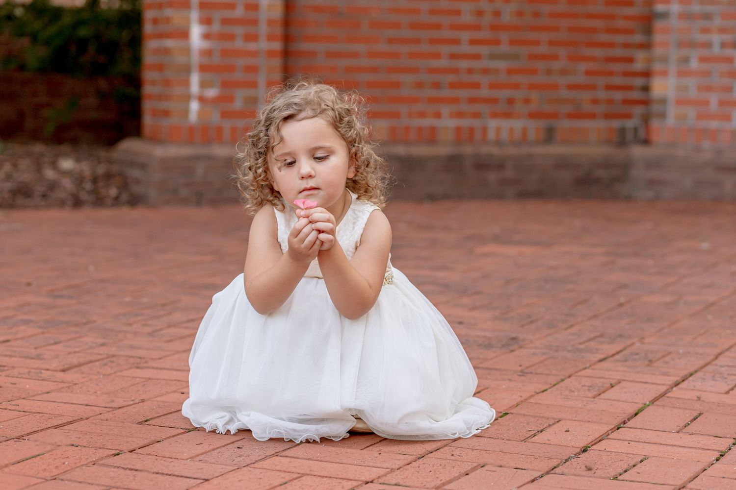 A curly-haired little girl in a white dress kneels on brick pavement with hands clasped in prayer.