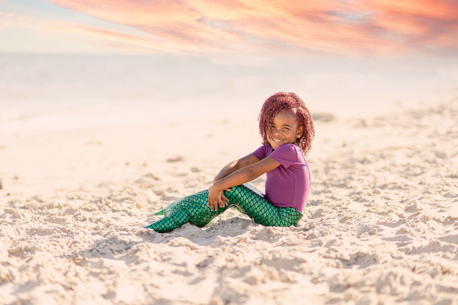 Young girl in mermaid costume sitting on sandy beach at colorful sunset.