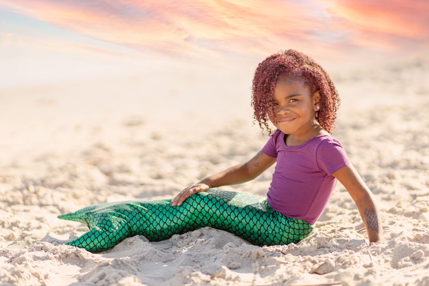 Smiling young girl wearing green mermaid tail costume on beach at sunset.