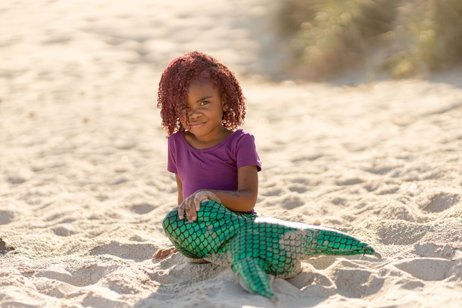 Young girl with curly red hair wearing green mermaid tail sits on beach.