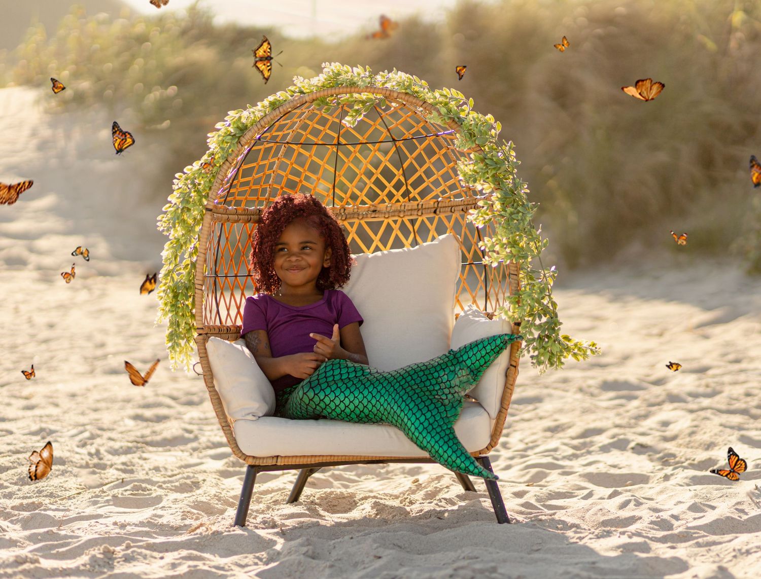 Young girl in mermaid costume sits in wicker chair on beach with butterflies.