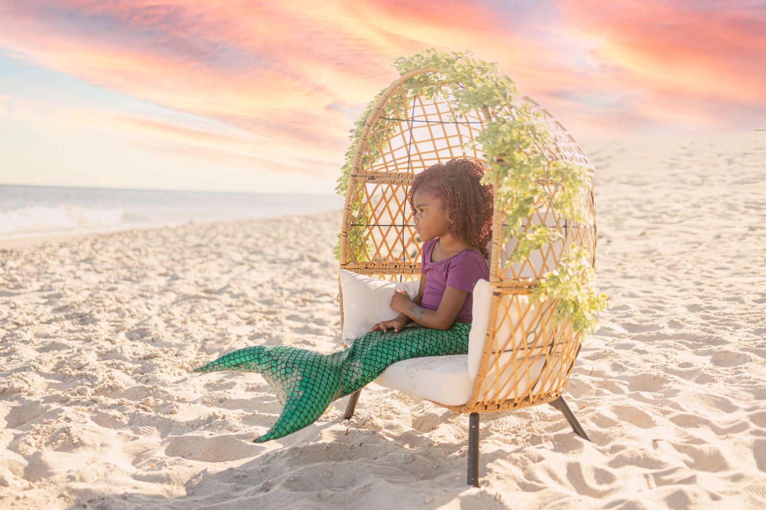 A young girl dressed as a mermaid sits in a wicker egg chair on a sandy beach at sunset.