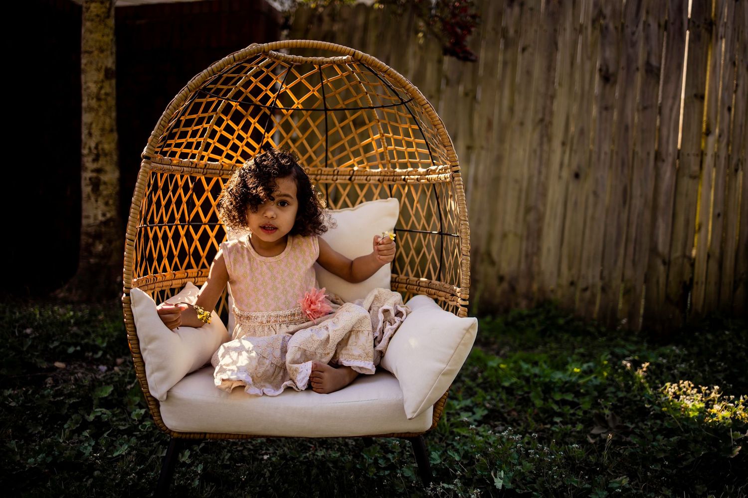 Young girl playing with a pillow in a wicker egg chair outdoors.