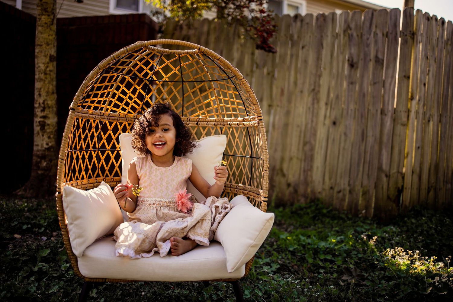 Happy toddler girl sitting in a wicker egg chair in a sunny backyard.