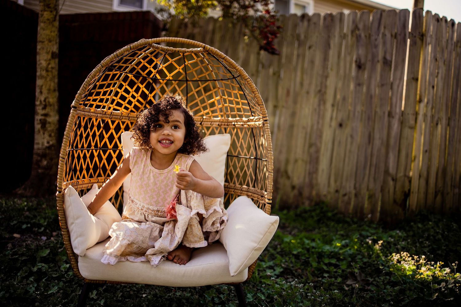 Smiling toddler girl sitting in a wicker egg chair in a backyard.